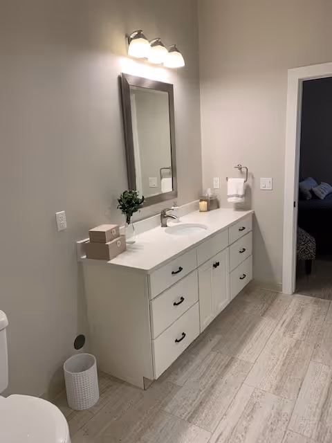 A clean and modern bathroom featuring a white vanity with multiple drawers and a sink, a rectangular mirror above the sink, a three-light fixture mounted on the wall, a small plant and decorative boxes on the countertop, a towel ring with a white towel, and a glimpse into an adjacent room with a bed and pillows.