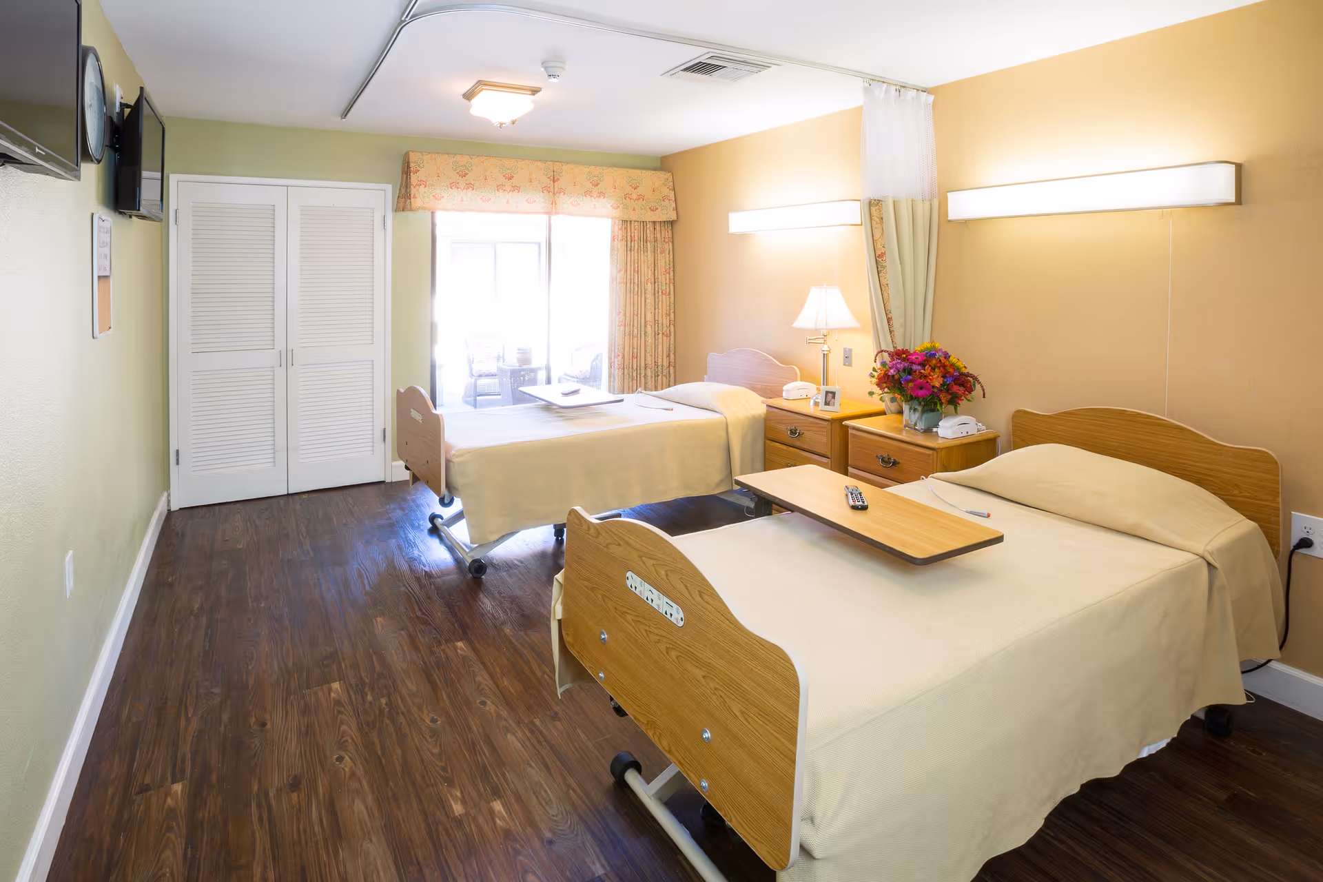 A bright and clean room with two hospital-style beds covered in beige blankets, each with an overbed table. There is a nightstand between the beds with a lamp, a telephone, and a vase of colorful flowers. The room has wooden flooring, a closet with white louvered doors, a wall-mounted TV, and a large window with floral curtains letting in natural light.