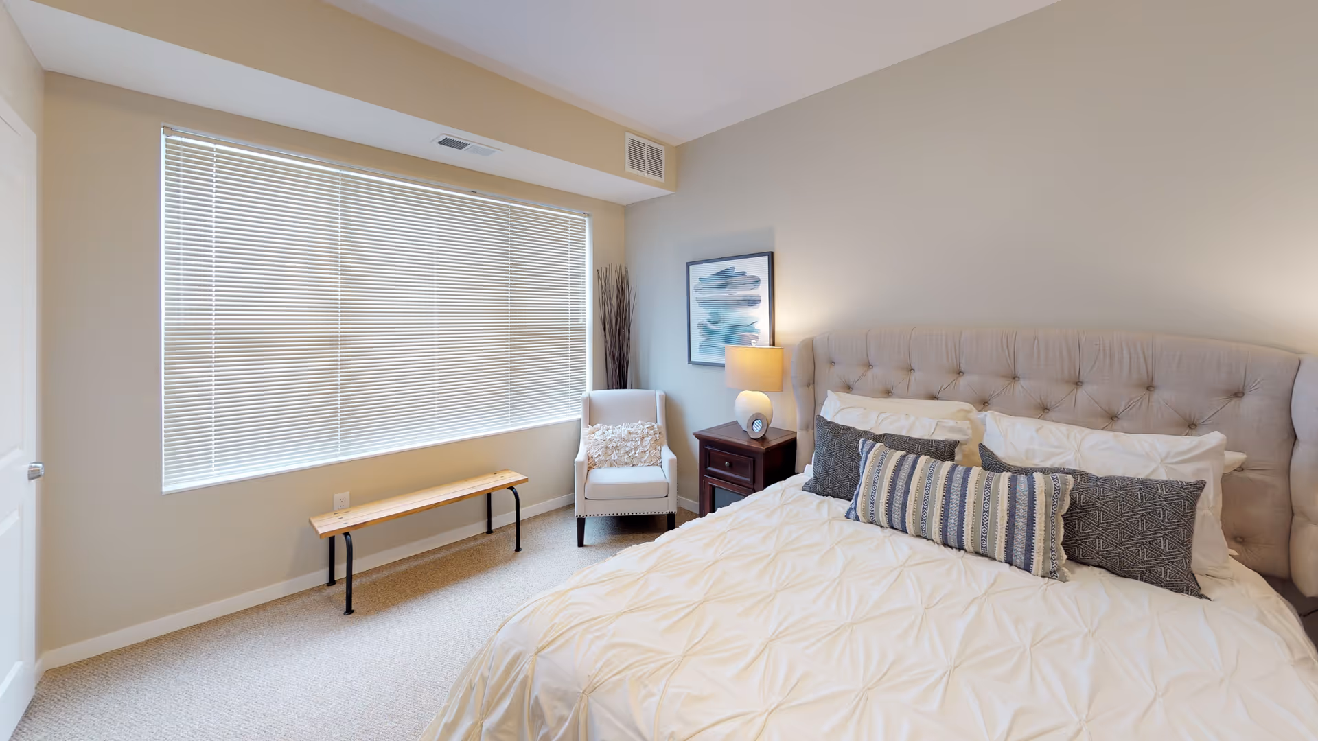 A bright and cozy bedroom featuring a large bed with a tufted headboard, white bedding, and multiple decorative pillows. Next to the bed is a wooden nightstand with a lamp and a framed abstract artwork on the wall. A white armchair with a textured pillow sits near a large window with closed blinds. A simple wooden bench is placed beneath the window. The room has beige walls and carpeted flooring.