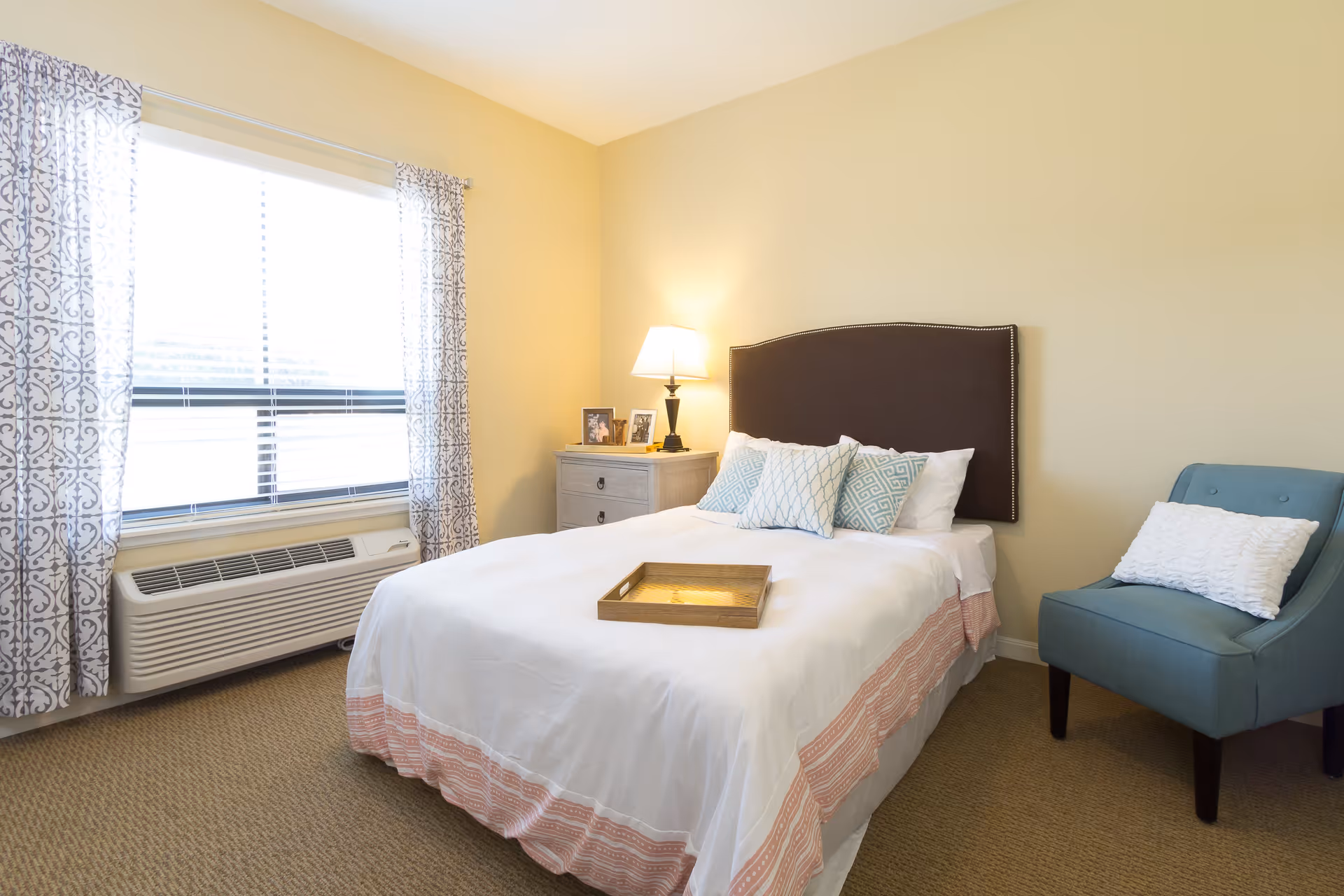 A bright bedroom with a large window covered by patterned curtains, a bed with white and pink bedding and multiple pillows, a wooden tray on the bed, a nightstand with a lamp and framed photos, and a blue upholstered chair with a white pillow.