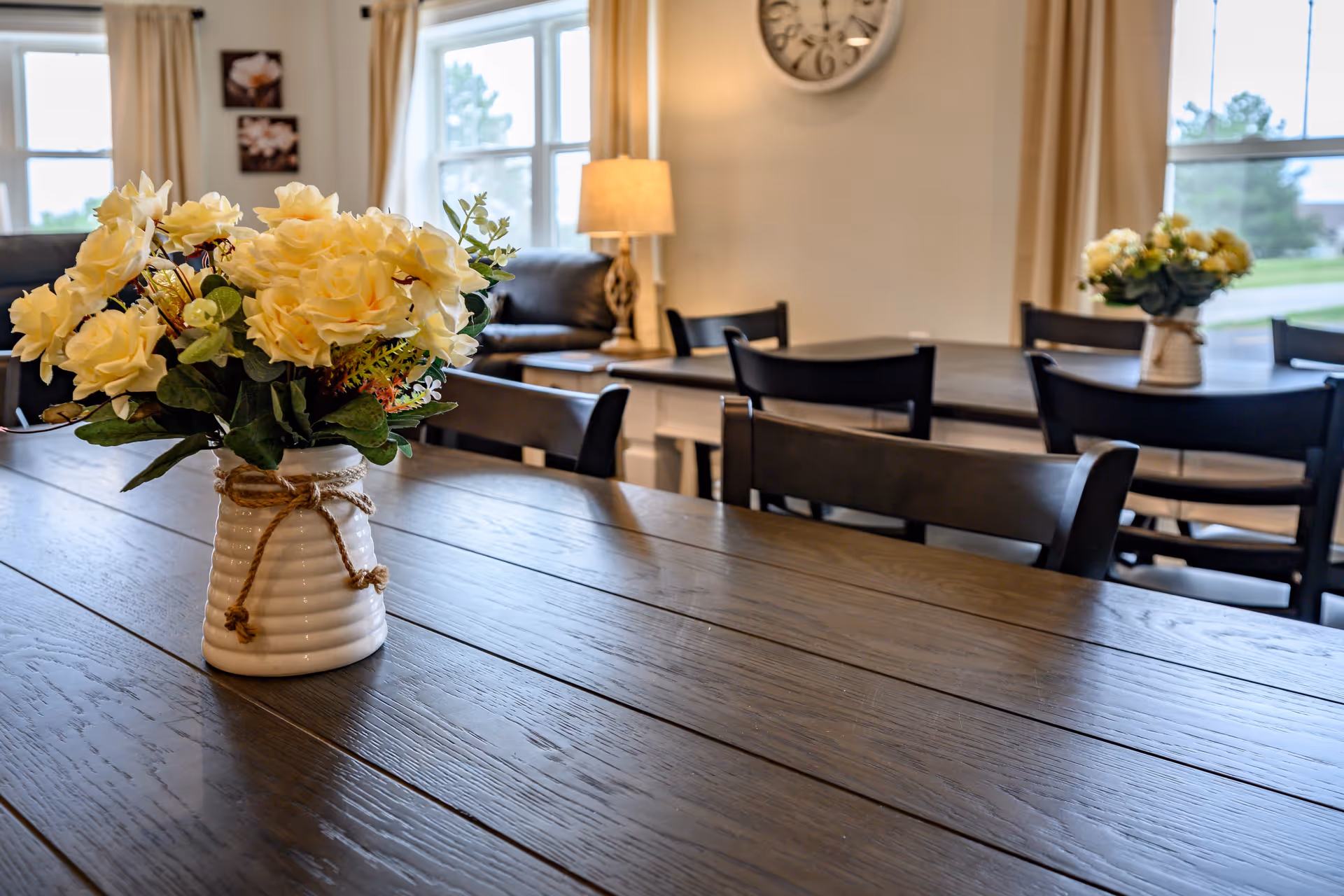 A dining room table with a vase of yellow flowers, surrounded by black chairs and a cozy seating area in the background, featuring large windows and soft lighting.