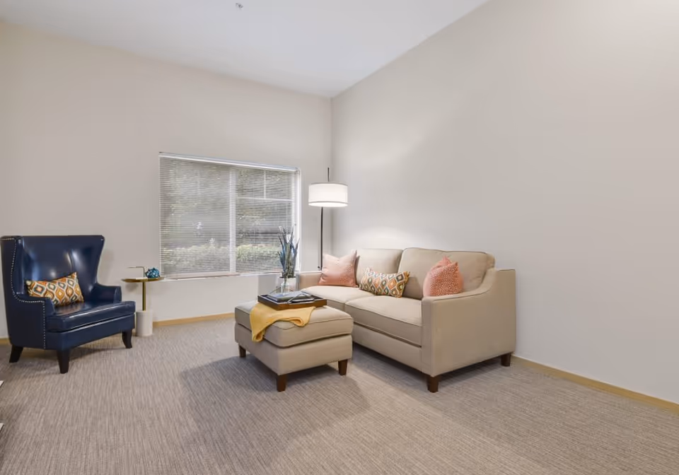 Bright living room with a beige sofa and ottoman, a blue armchair, floor lamp, and a window.