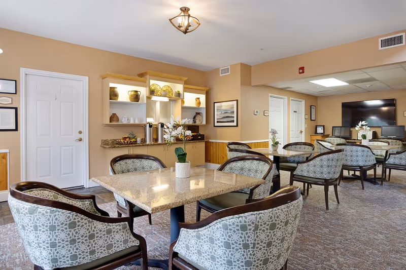 A well-lit common area with multiple square tables surrounded by patterned upholstered chairs. The room has beige walls, a carpeted floor, and a small counter with decorative pottery and plates on shelves. There is a large flat-screen TV mounted on the far wall and framed artwork on the walls.