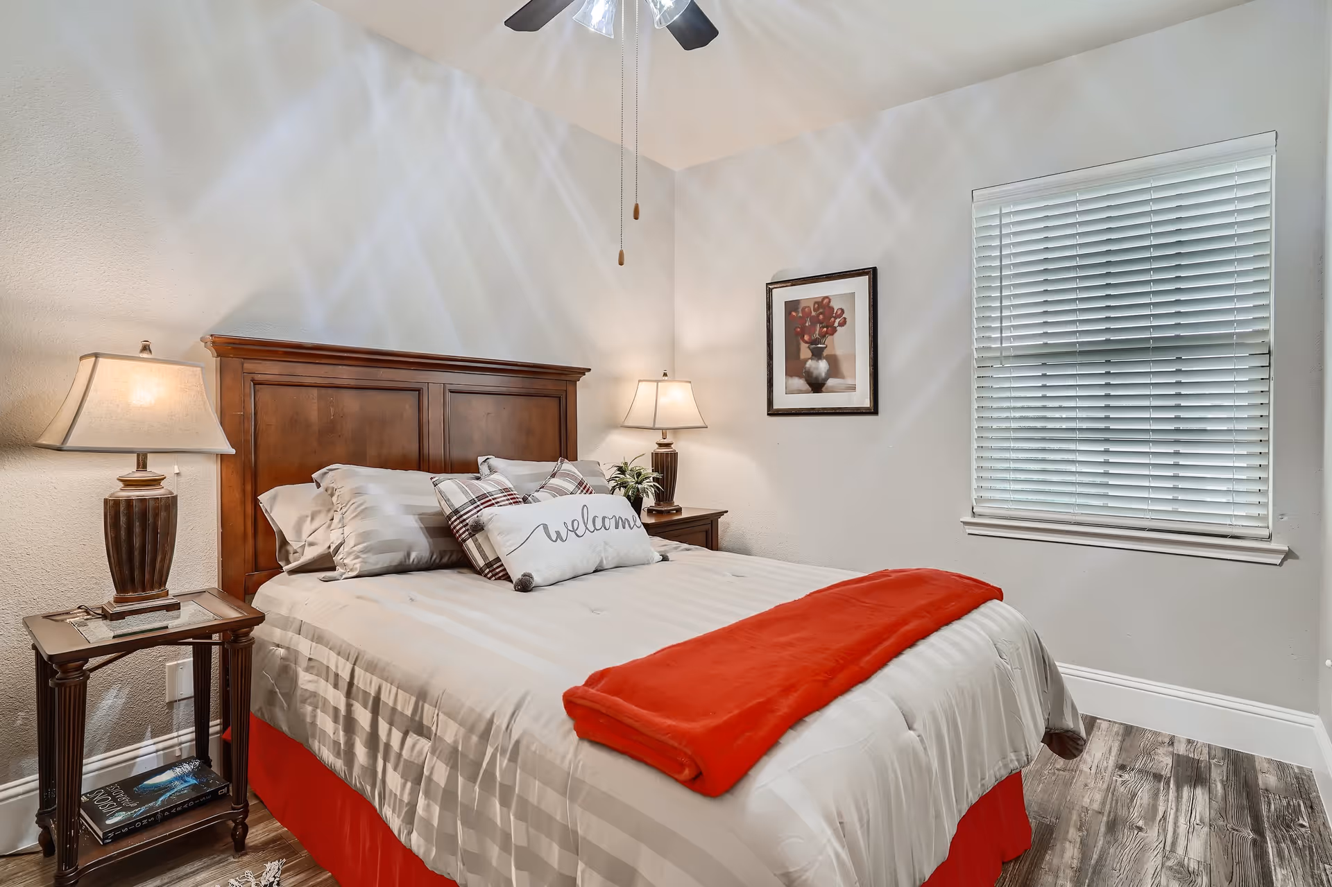 A cozy bedroom with a wooden headboard bed dressed in white and beige striped bedding, a red throw blanket, and decorative pillows including one with the word 'welcome'. There are two bedside tables with lamps, a framed floral artwork on the wall, a window with white blinds, and a ceiling fan above.
