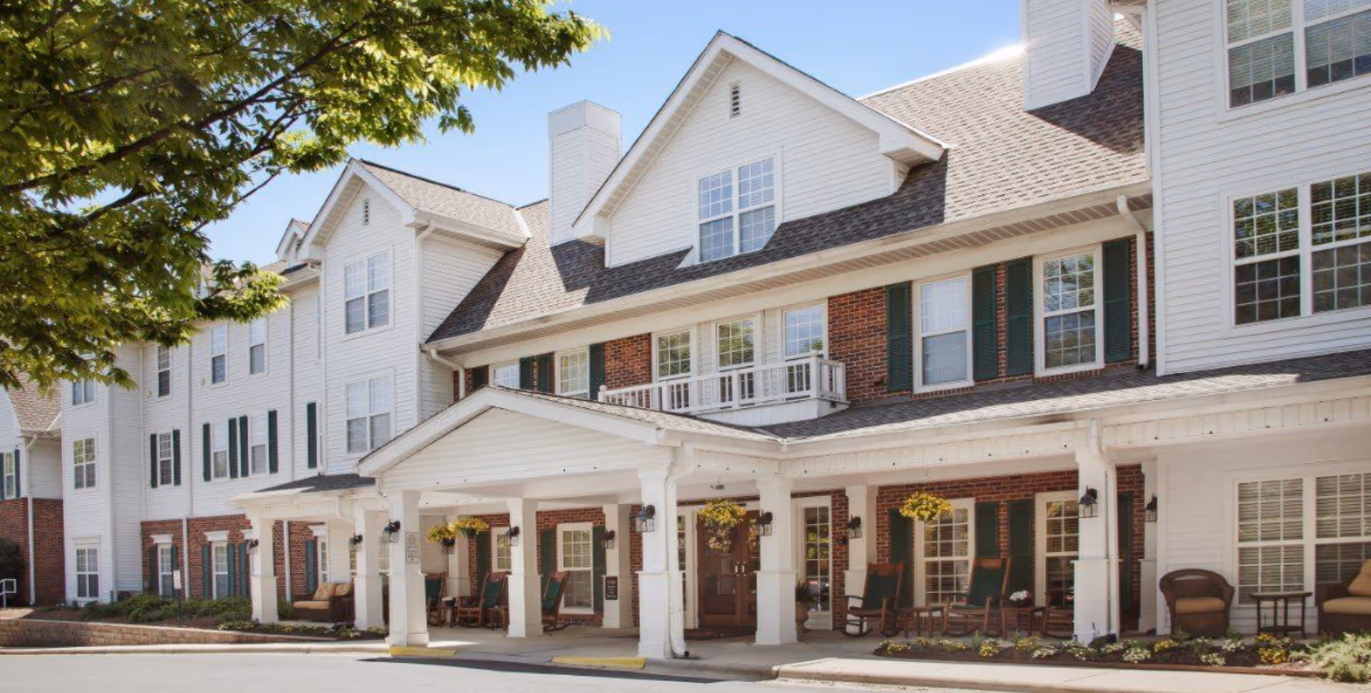 Front exterior of a multi-story senior living facility with a covered porte-cochere, rocking chairs on the porch, and hanging plants.