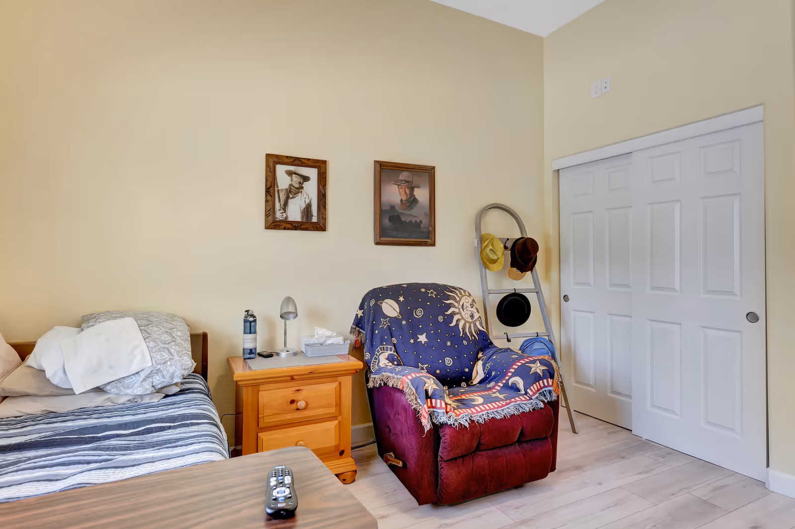 A cozy senior living bedroom with a bed on the left side covered with striped bedding and pillows. Next to the bed is a wooden nightstand with a lamp, water bottle, and tissue box. A comfortable maroon recliner chair draped with a celestial-themed blanket is positioned beside the nightstand. On the wall above the chair are two framed pictures of men wearing hats. A wooden ladder leaning against the wall holds several hats. There is a white sliding closet door on the right side of the room. The floor is light-colored wood, and the walls are painted a soft beige.