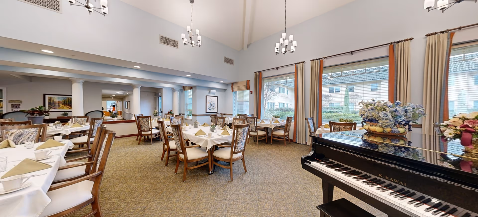 A spacious dining room with multiple round tables covered with white tablecloths, each set with folded napkins and glassware. Wooden chairs with cushioned seats surround the tables. Large windows with curtains allow natural light to fill the room. A black grand piano with floral arrangements on top is visible in the foreground on the right side. The room has high ceilings with hanging light fixtures and a carpeted floor.