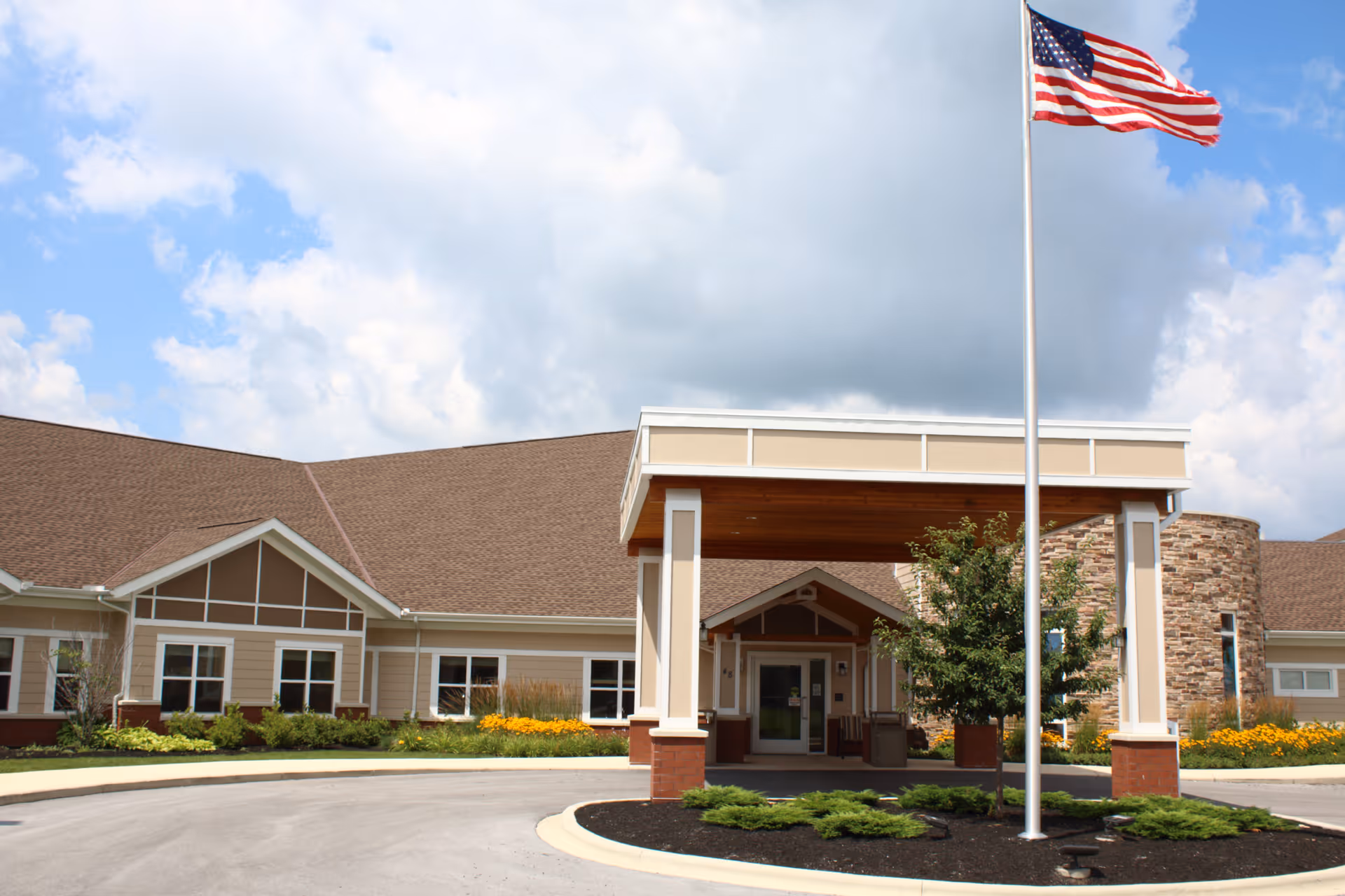 Exterior view of Tiffin Rehabilitation Center showing the entrance with a covered drop-off area, a flagpole with an American flag, landscaped greenery, and a partly cloudy sky.