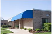Exterior view of a single-story brick building with a blue awning over the entrance, surrounded by green shrubs and a concrete walkway.