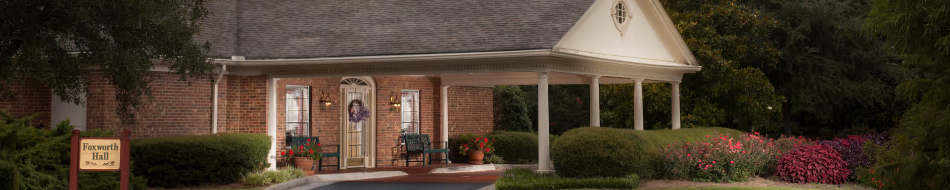 Exterior view of a brick building with a covered entrance supported by white columns, surrounded by well-maintained bushes and colorful flowers, with a sign reading 'Foxworth Hall' near the entrance.