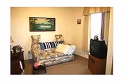 Small living room with a patterned loveseat, side tables, a TV on a stand, a framed landscape painting on the wall, and a curtained window.