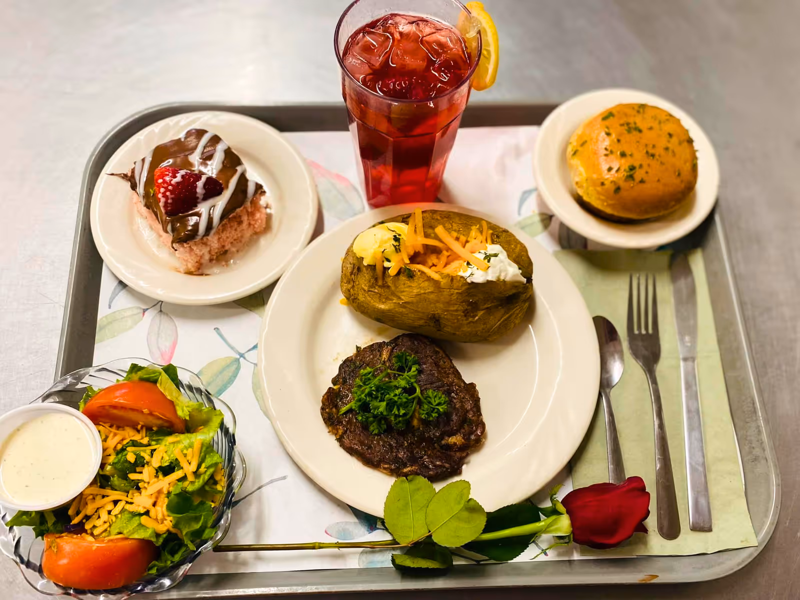 A meal tray with a baked potato topped with butter, shredded cheese, and sour cream, a piece of cooked meat garnished with parsley, a side salad with lettuce, tomato, shredded cheese, and a cup of dressing, a bread roll, a dessert with chocolate and strawberry topping, and a glass of iced tea with a lemon slice. A red rose is placed on the tray next to the utensils.