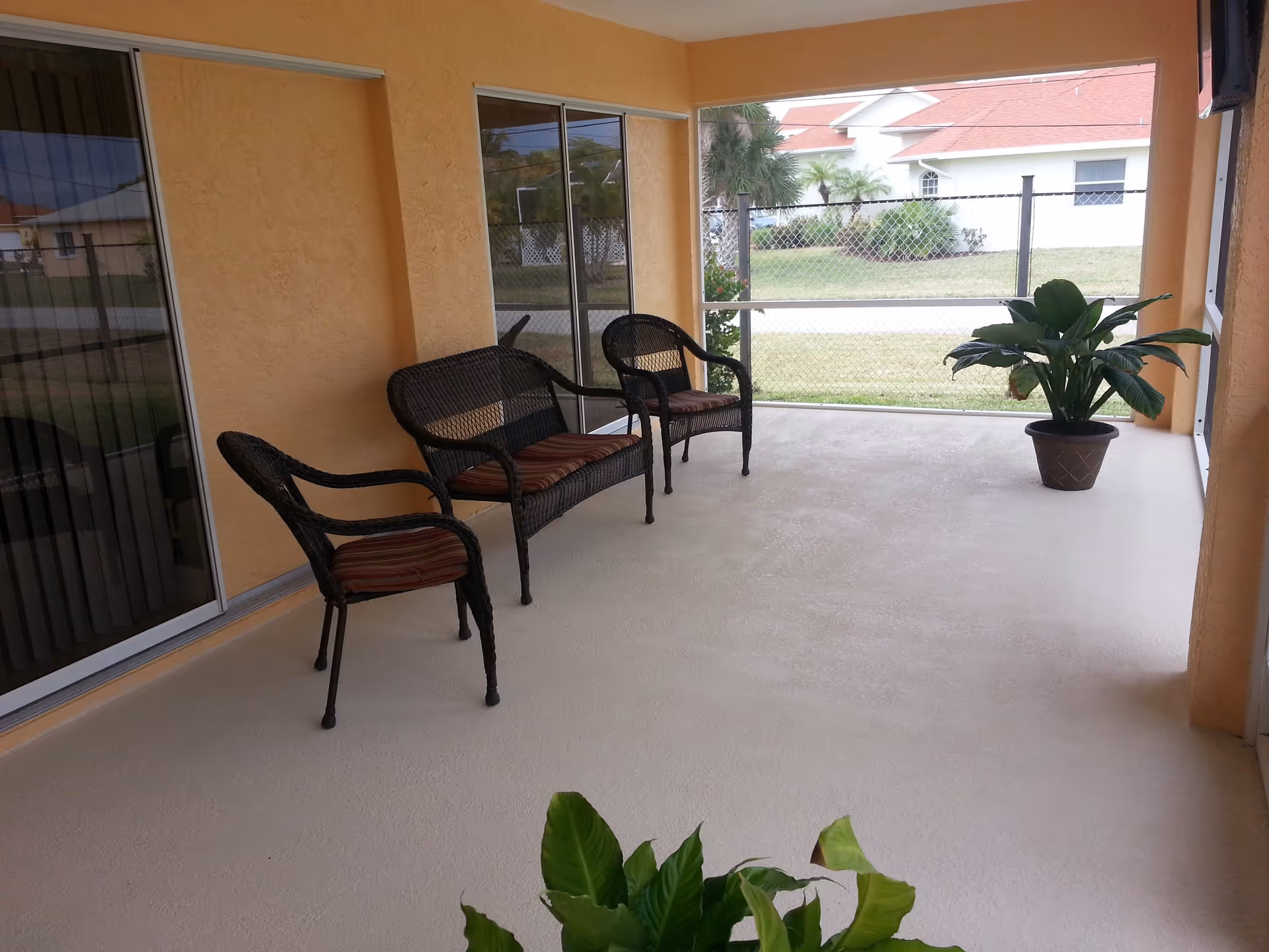 A covered outdoor patio area with three wicker chairs with striped cushions arranged along a peach-colored wall. There are two potted plants on the patio floor, and a chain-link fence with a view of a grassy yard and neighboring houses with red roofs in the background.