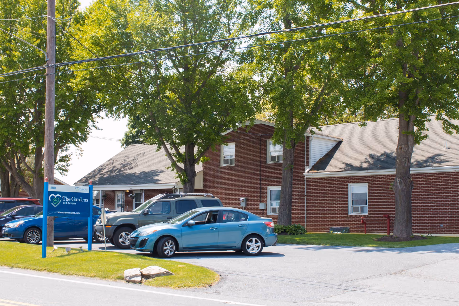 Exterior view of The Gardens at Stevens senior living facility showing a brick building with several windows, a parking area with multiple cars, green trees, and a blue sign with the facility's name and website.
