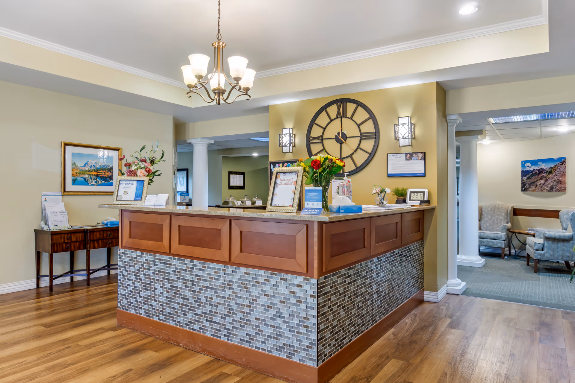 Reception area of Brookdale Olympia East featuring a wooden front desk with mosaic tile accents, a large round wall clock, a chandelier, and floral arrangements. The space has wooden flooring, beige walls, and a seating area with armchairs visible in the background.
