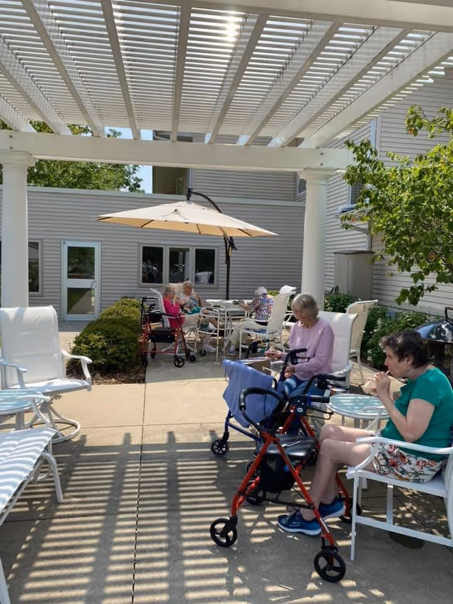 Outdoor patio area at a senior living facility with several elderly women sitting on white chairs around tables under a large umbrella and a pergola. Some women use walkers and wheelchairs. The area is surrounded by bushes and the building exterior is visible in the background.