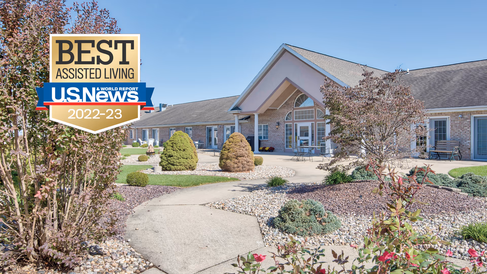Exterior view of The Villas at St. James, showing a single-story building with a peaked roof, large windows, and a landscaped garden with shrubs, trees, and a paved walkway under a clear blue sky.