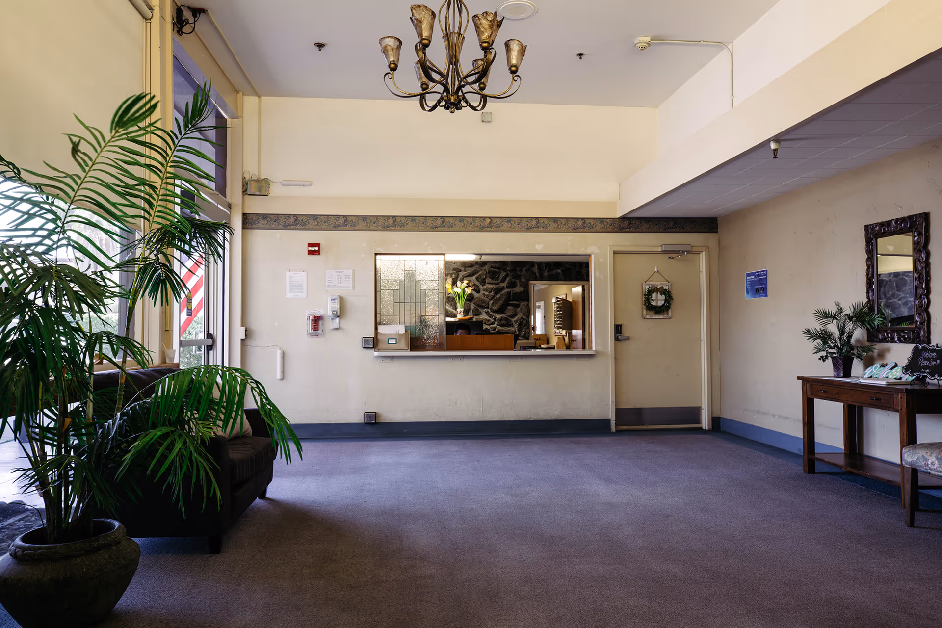 A spacious lobby area with a large potted plant on the left, a dark armchair near the glass entrance door, a chandelier hanging from the ceiling, and a reception window with a stone wall visible behind it. On the right side, there is a wooden table with plants and a mirror above it, and a door with a wreath hanging on it.