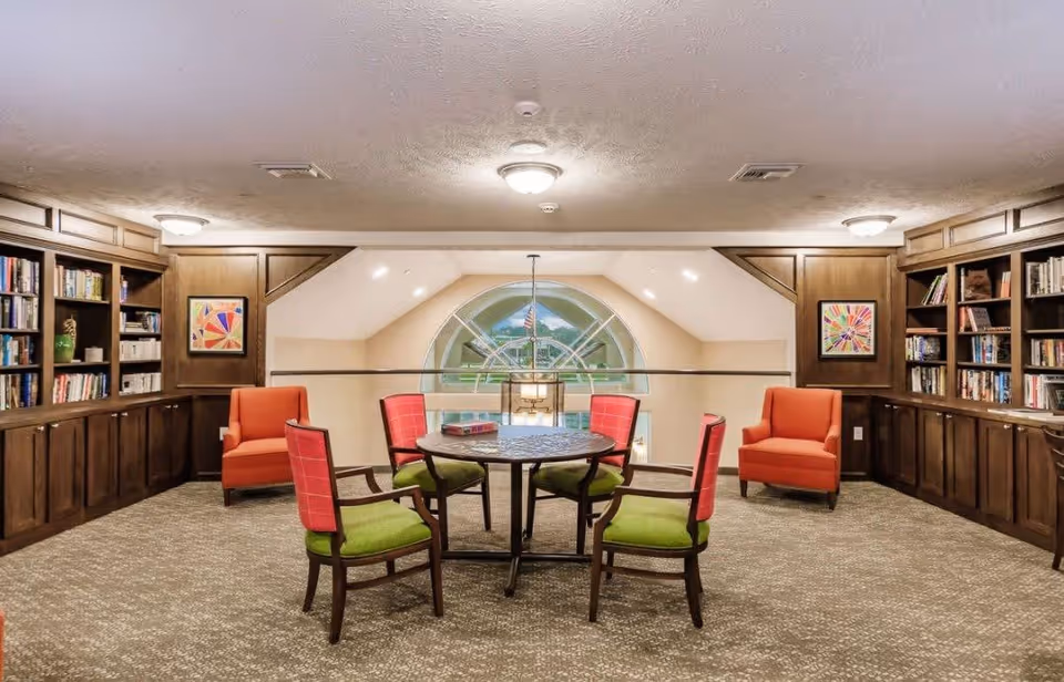 A cozy library or reading room with built-in dark wood bookshelves filled with books on both sides. In the center, there is a round table with four chairs featuring green seats and red backs. Two orange armchairs are placed against the back wall. A large arched window with a view of an American flag outside is visible in the background. The room has a carpeted floor and ceiling lights.
