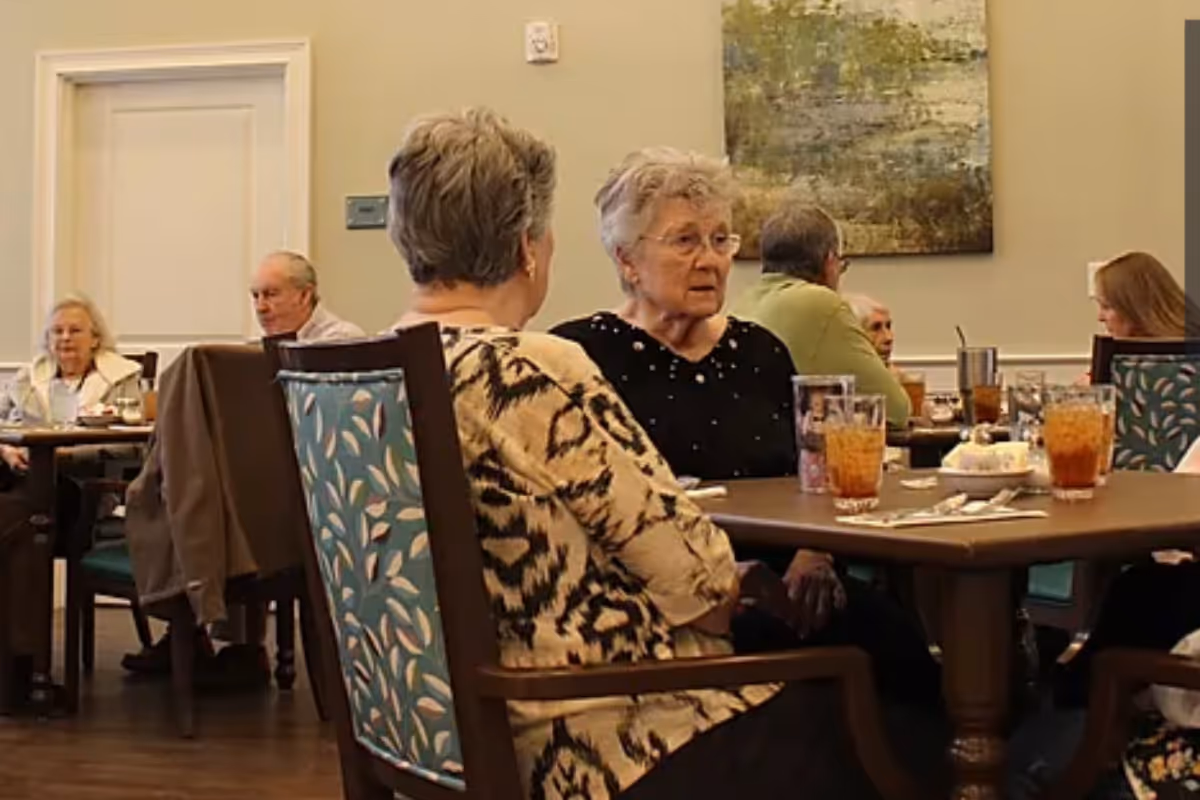 Several elderly people sitting at tables in a dining room, engaged in conversation and having drinks. The room has light-colored walls, a large painting, and wooden chairs with patterned upholstery.