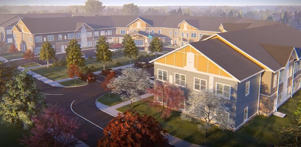 Aerial view of a senior living facility named Harmony at Anderson, showing multiple connected buildings with beige and gray siding, surrounded by landscaped grounds with trees and parking spaces. The scene is set in a suburban area with a forested background under a clear sky.