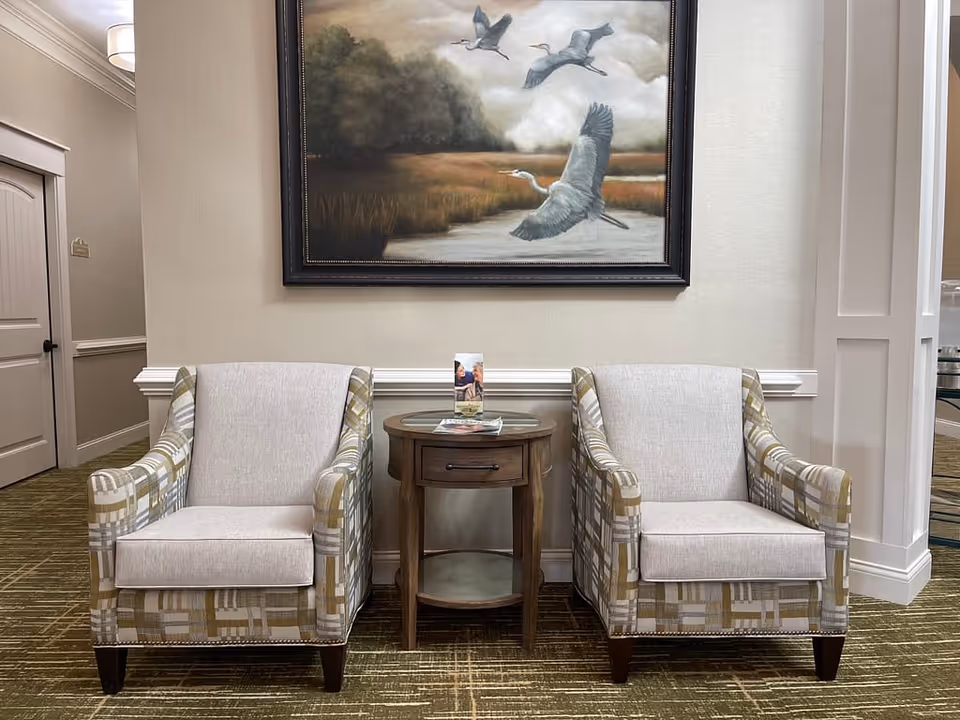 Two patterned armchairs with light beige cushions are placed on either side of a small round wooden table with a drawer and a glass top. On the table are some brochures. Above the chairs hangs a large framed painting depicting three flying herons over a marshy landscape. The setting appears to be a quiet, well-lit interior space with beige walls and carpeted floor.