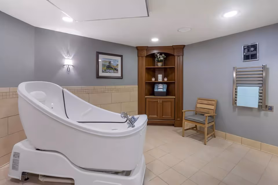 A spacious bathroom with a large white assisted bathing tub, beige tiled walls and floor, a wooden chair with a cushion, a wooden cabinet with shelves holding towels and decorative items, a wall-mounted towel rack with a blue towel, and a framed picture on the gray wall.