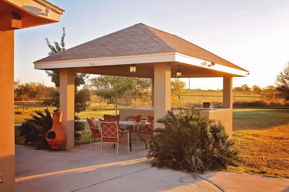 Covered outdoor patio gazebo with a dining table and chairs on a concrete slab in a grassy yard at sunset.