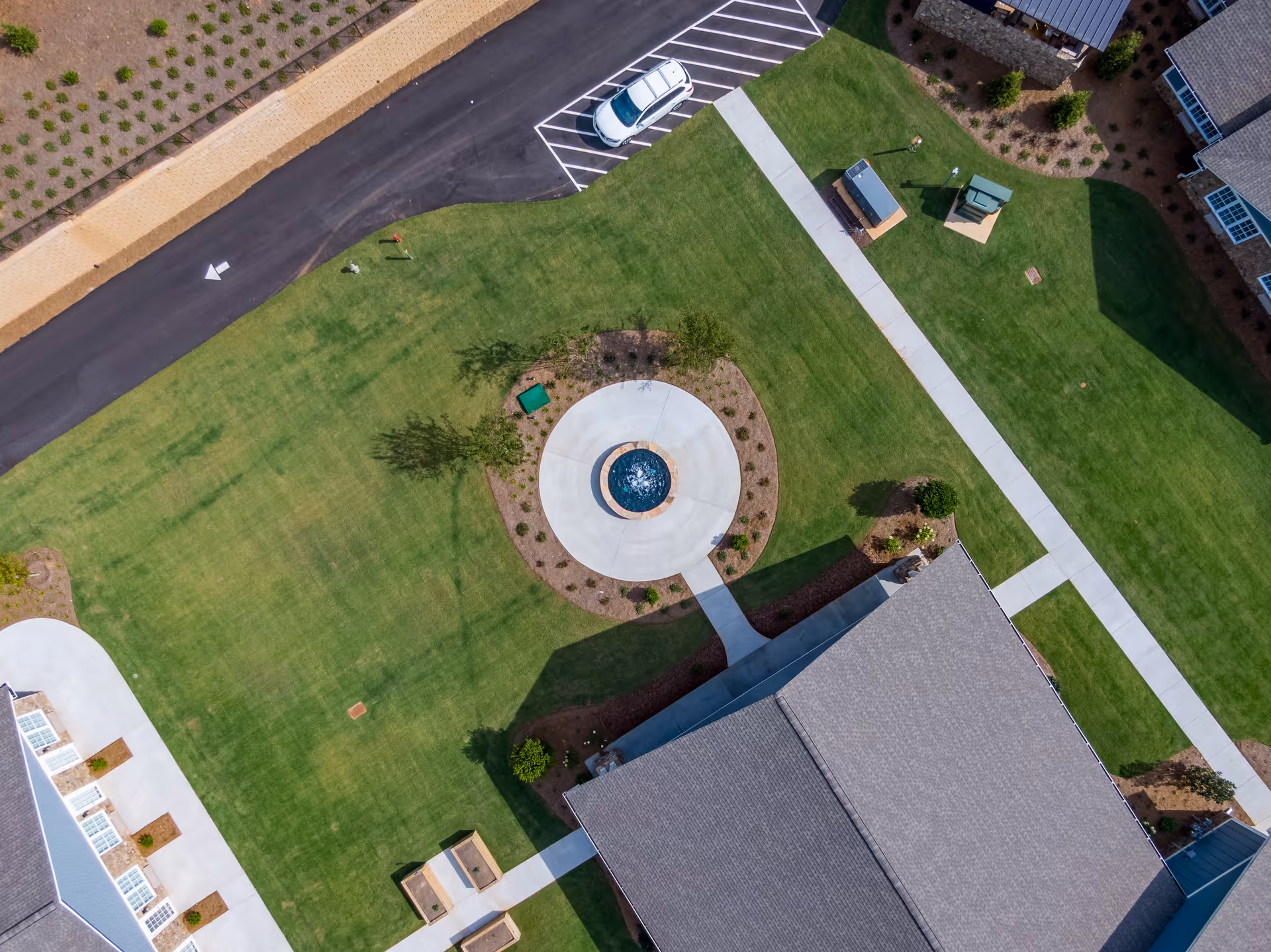 Aerial view of a landscaped outdoor area at Manor Lake Assisted Living & Memory Care - Hiram, featuring a circular fountain surrounded by a concrete walkway, green lawns, a parking area with a white car, and parts of buildings with gray roofs.