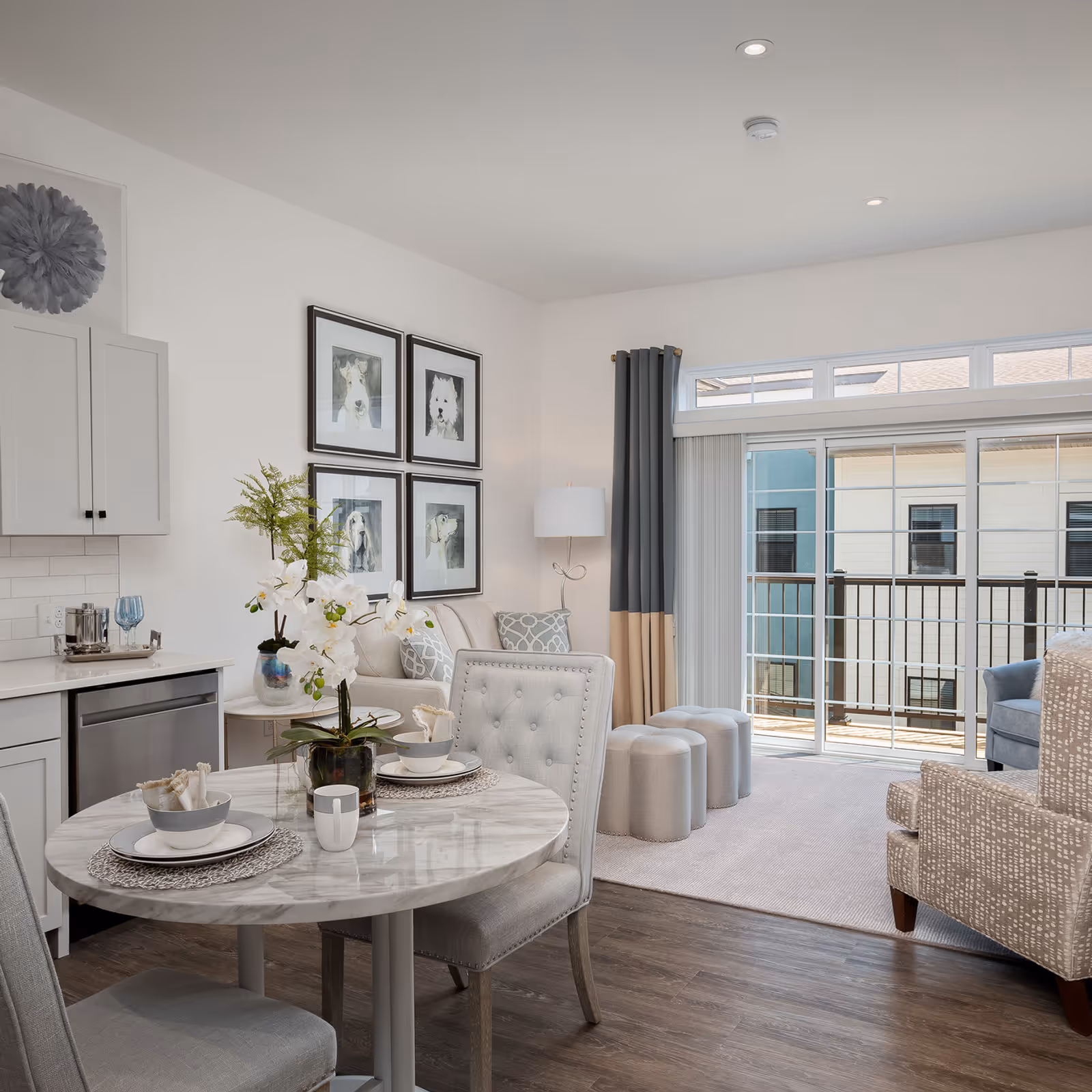 A bright and modern senior living apartment interior featuring a small round dining table set for two with gray upholstered chairs, a kitchenette with white cabinets and stainless steel dishwasher, a cozy living area with a beige sofa, patterned armchair, and three small ottomans. Large sliding glass doors with curtains open to a balcony with a view of neighboring buildings.