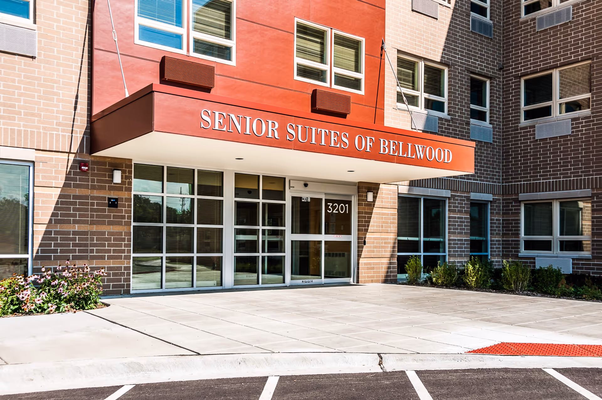 Entrance of Senior Suites of Bellwood building with a red canopy displaying the facility name above glass doors and windows. The building exterior is made of brick with several windows and some landscaping with flowers and shrubs near the entrance.