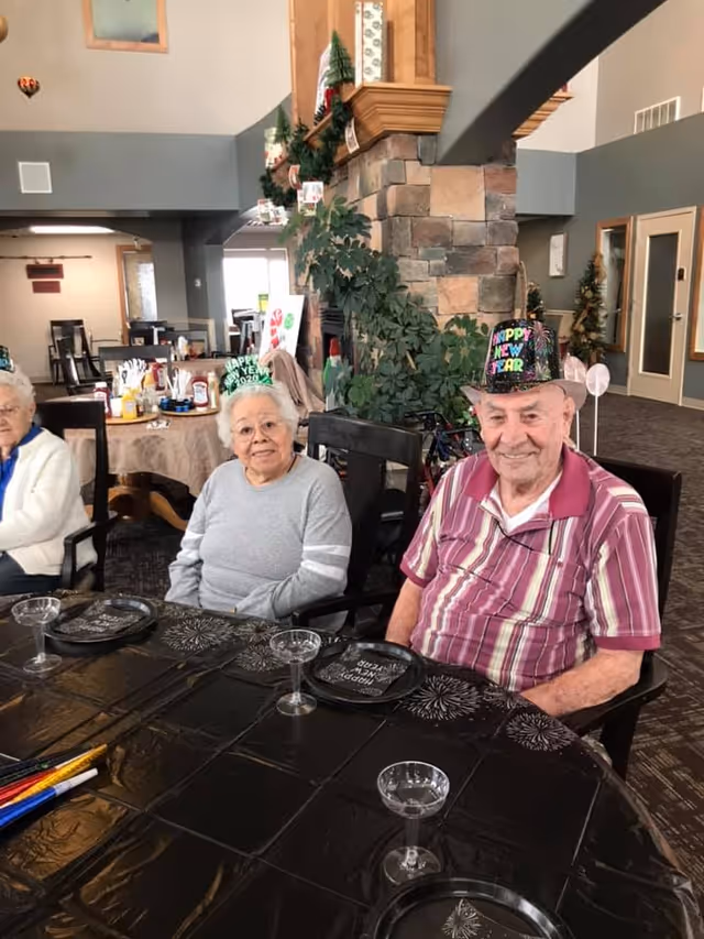 Three elderly residents wearing New Year's hats sit at a decorated dining table in a senior living communal room.