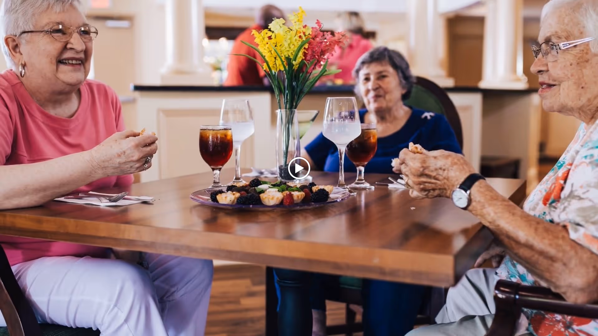 Three elderly women sitting around a wooden dining table in a well-lit room, enjoying snacks and drinks. The table has a vase with yellow and red flowers and a plate with assorted small desserts. The women appear to be engaged in conversation and smiling.