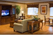 A cozy living room area with a green sofa, wooden coffee table, and a wooden entertainment center with a television. There is a potted plant next to the entertainment center, a table lamp on a side table, and framed artwork on the wall. Two windows with valances allow natural light into the room. A dining table with chairs is visible in the background.