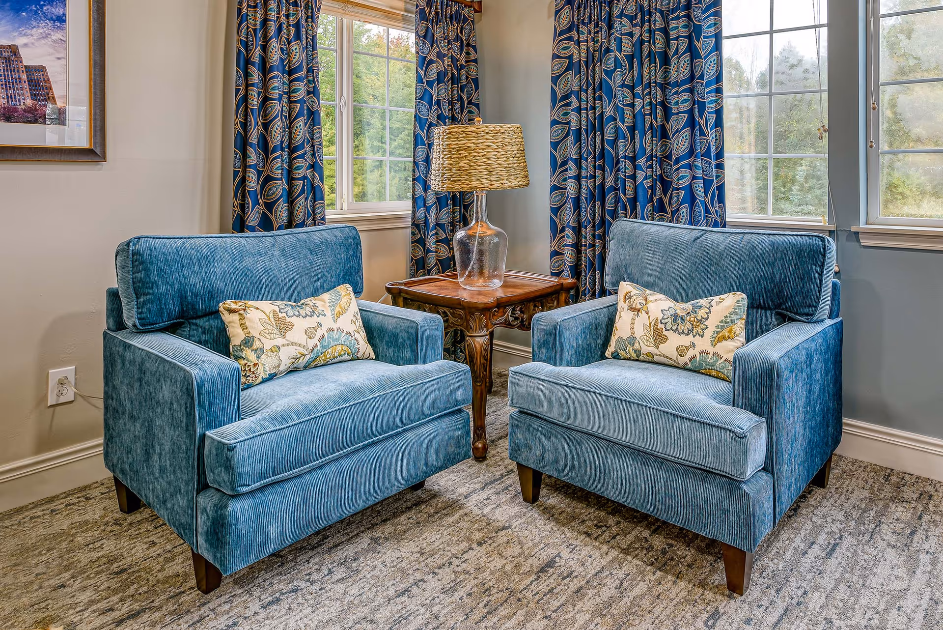 Two blue upholstered armchairs with floral patterned cushions are positioned facing each other with a wooden side table and a glass lamp with a woven lampshade between them. The room has large windows with blue curtains featuring a leaf pattern, and a framed picture on the wall.