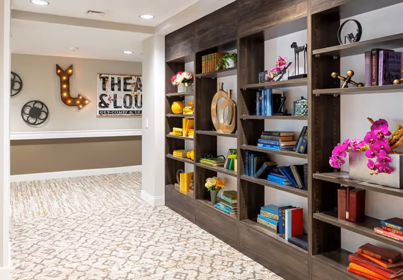 Carpeted common area with built-in bookshelves filled with books and decorative items and wall signage pointing toward a theatre/lounge.