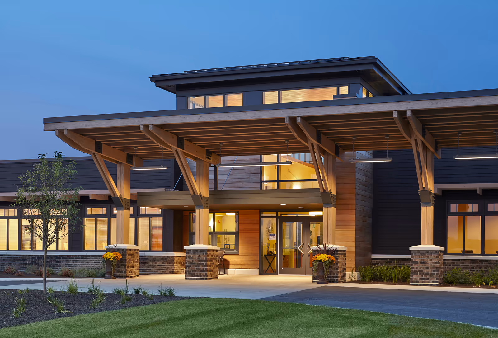 Entrance of a modern senior living facility with illuminated glass doors and a large wooden overhang at dusk