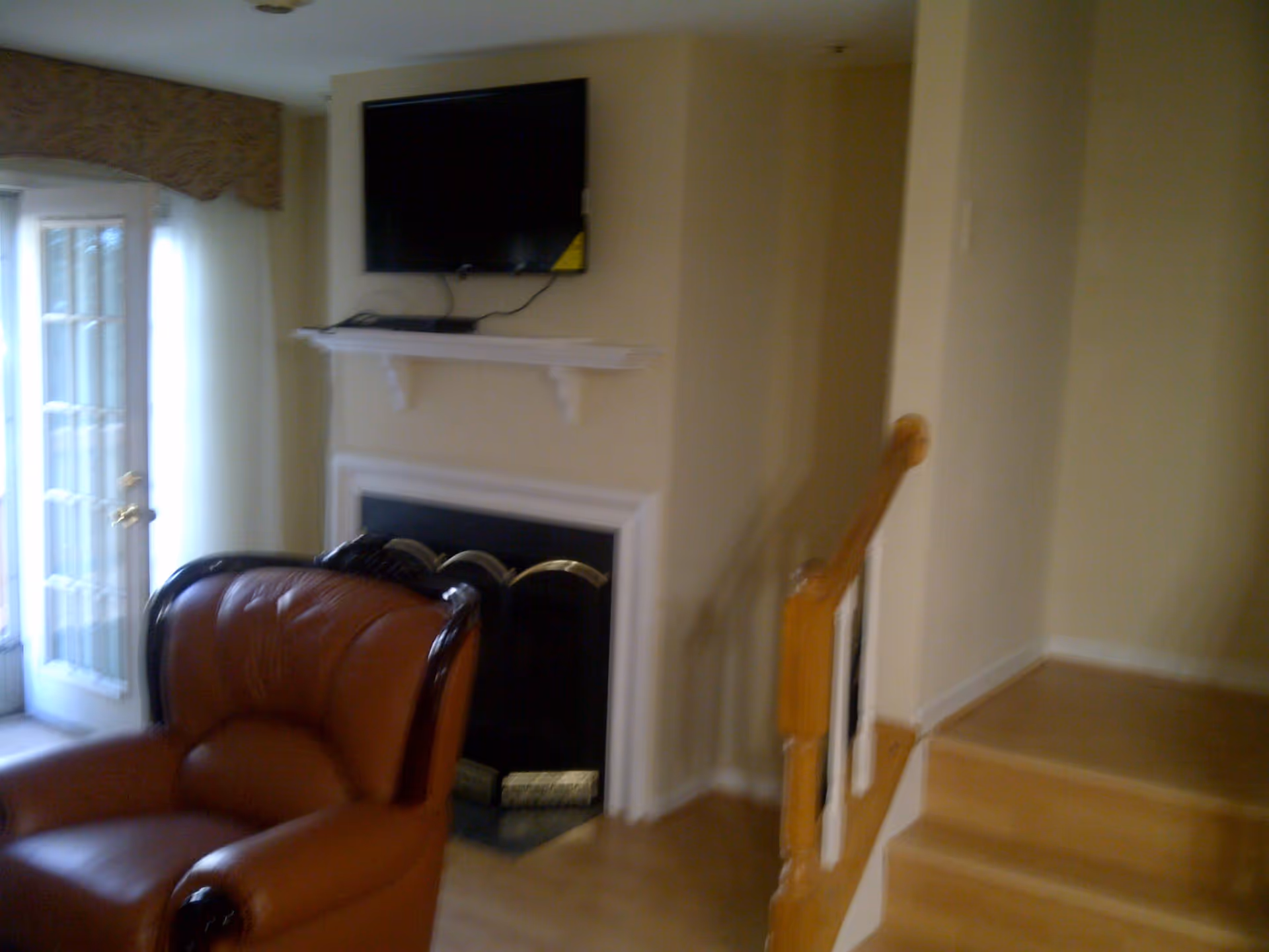 Interior living room with a leather armchair, a fireplace topped by a wall-mounted TV, stairs, and a glass door.