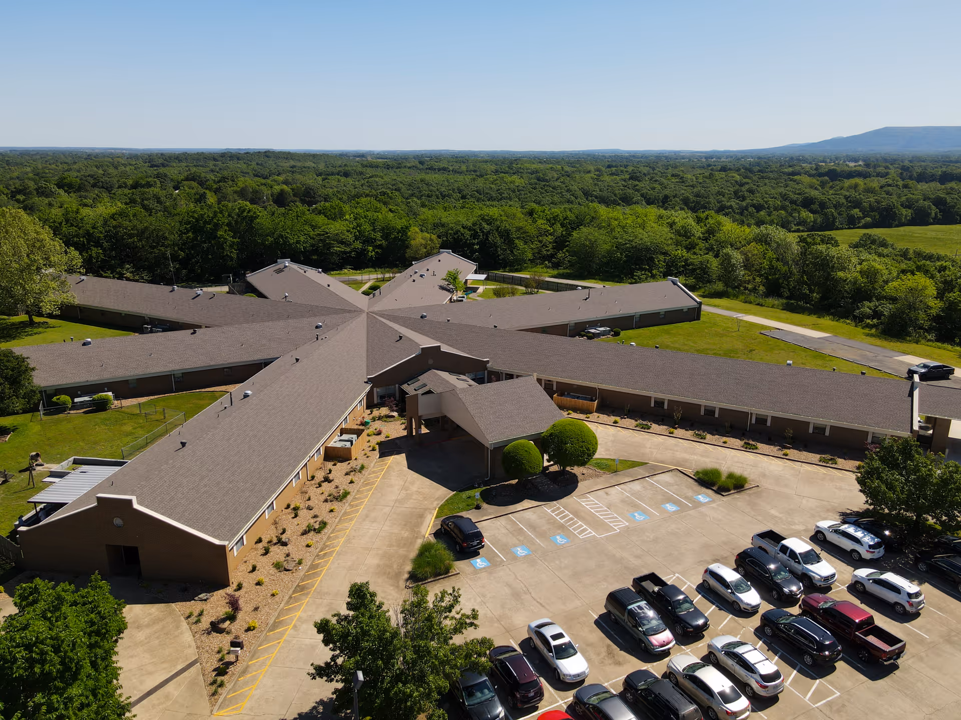 Aerial view of Oaks Healthcare Center showing a large, single-story building with multiple wings extending from a central entrance area. The building is surrounded by green trees and open land, with a parking lot filled with cars in the foreground under a clear blue sky.