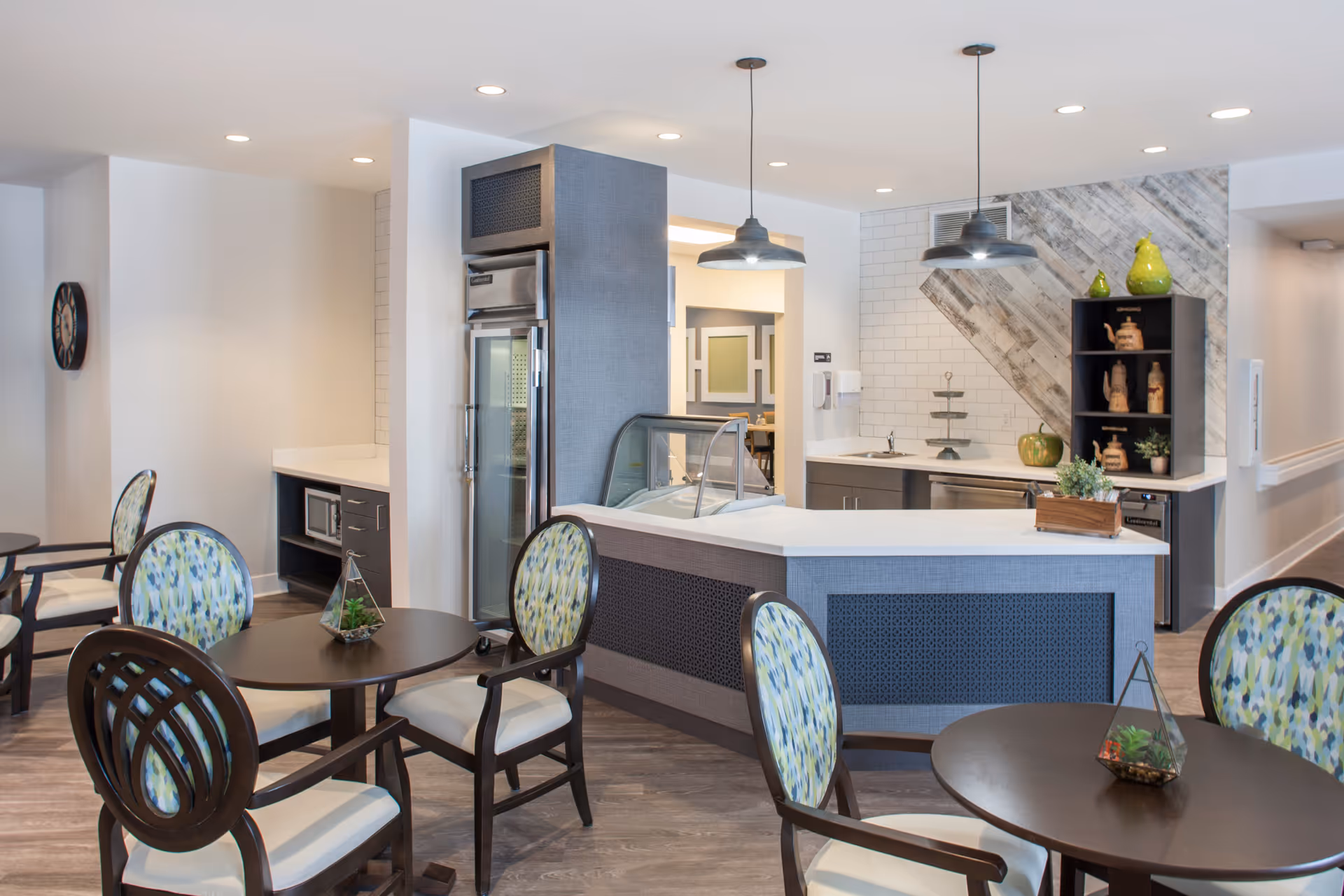 A modern dining area in a senior living facility featuring round tables with patterned cushioned chairs. The space includes a counter with a glass display case, pendant lighting, and a small kitchenette with a sink, cabinets, and decorative items on shelves. The flooring is wood-style, and the walls are light-colored with some decorative tiling.