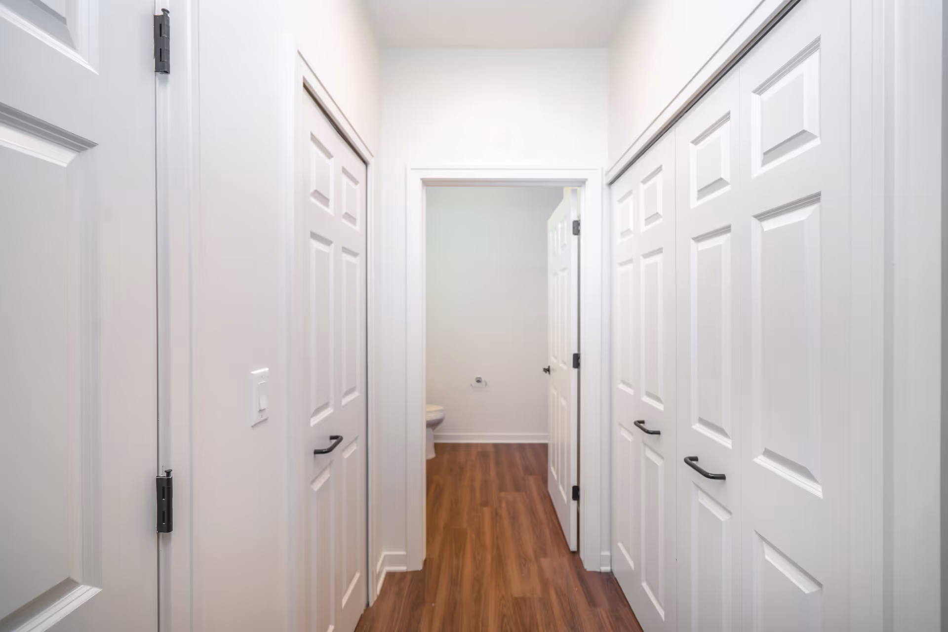 A bright interior hallway with white paneled doors and wood flooring leading to a small bathroom with a visible toilet.