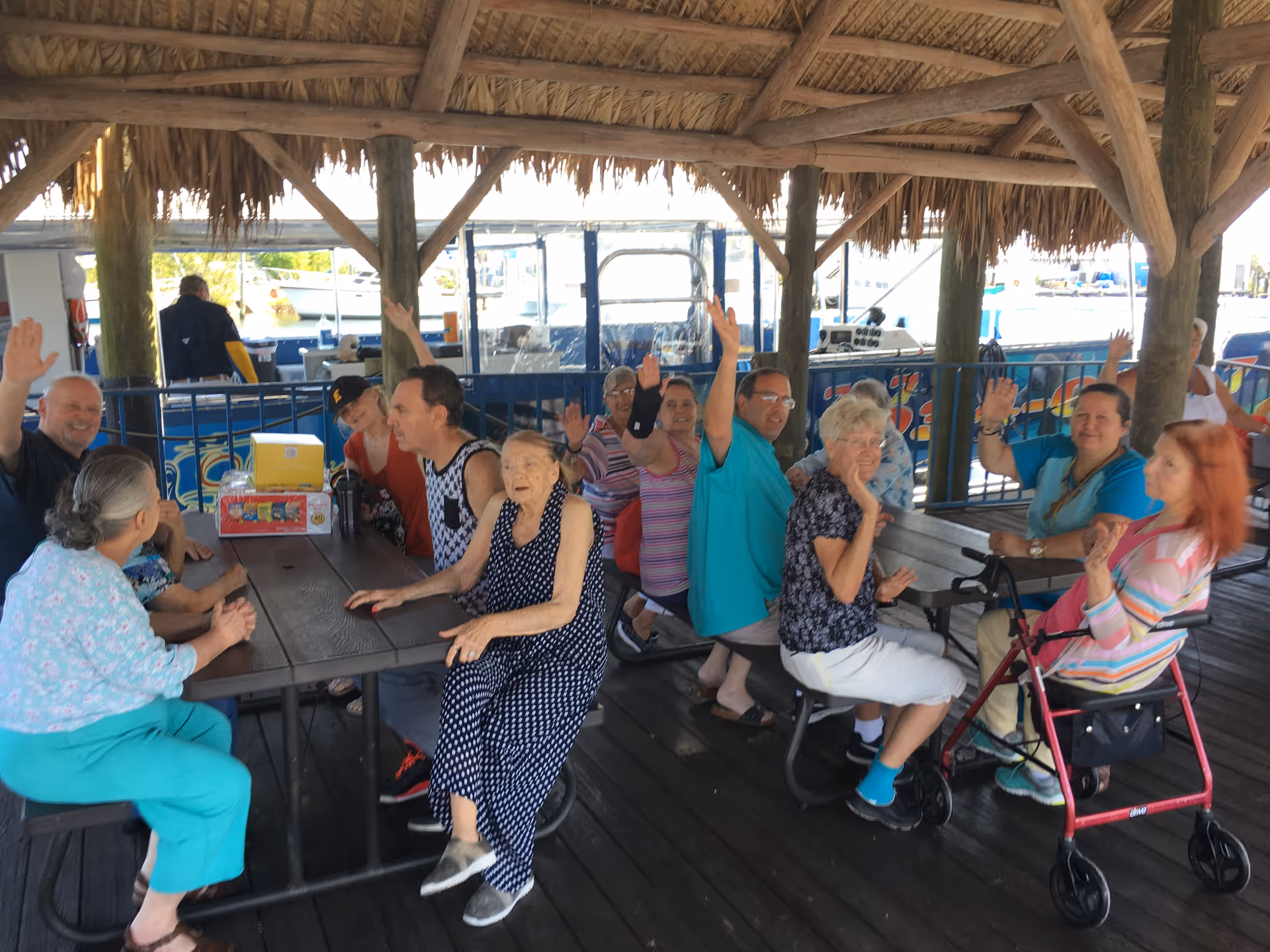 A group of elderly people sitting around a wooden picnic table under a thatched roof pavilion near a waterfront. Several individuals are raising their hands and smiling, with boats visible in the background.