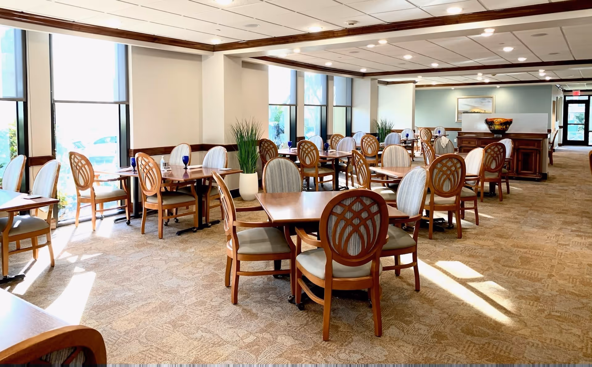 Bright dining room with multiple wooden tables and upholstered chairs arranged near large windows and potted plants.