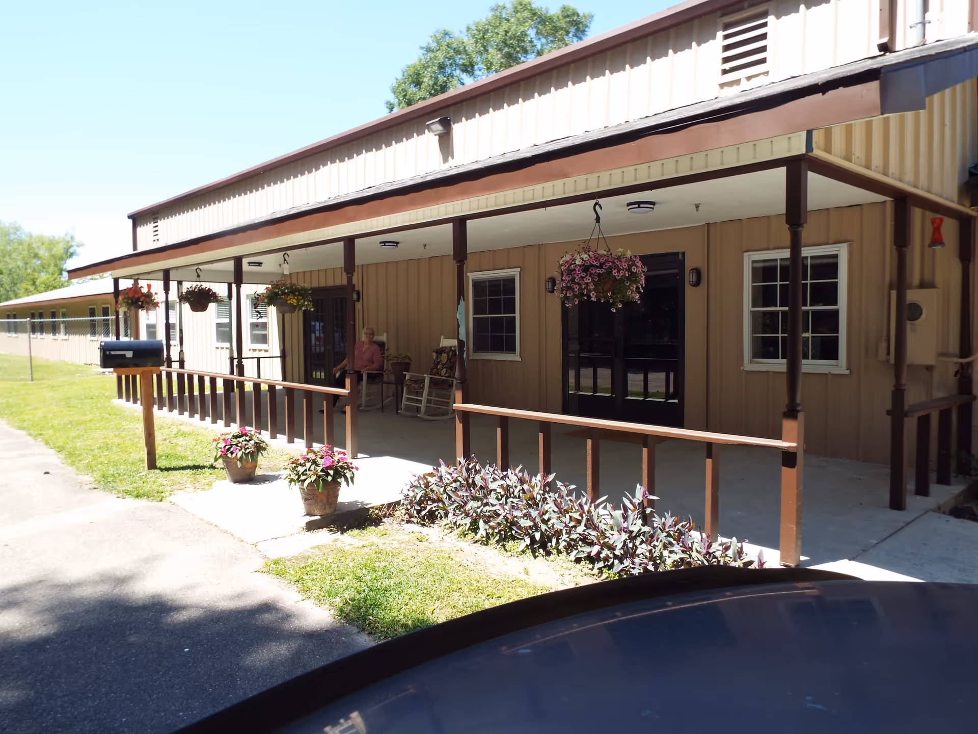 Exterior view of a single-story building with a covered porch featuring hanging flower baskets and potted plants. A person is sitting on a chair on the porch. The building has beige siding and several windows, with a clear blue sky in the background.