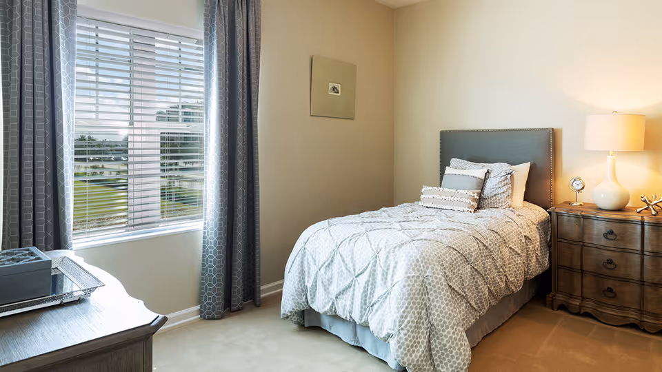 Sunlit bedroom with a twin bed dressed in patterned bedding, a wooden nightstand with a lamp, and a window with blinds and curtains.
