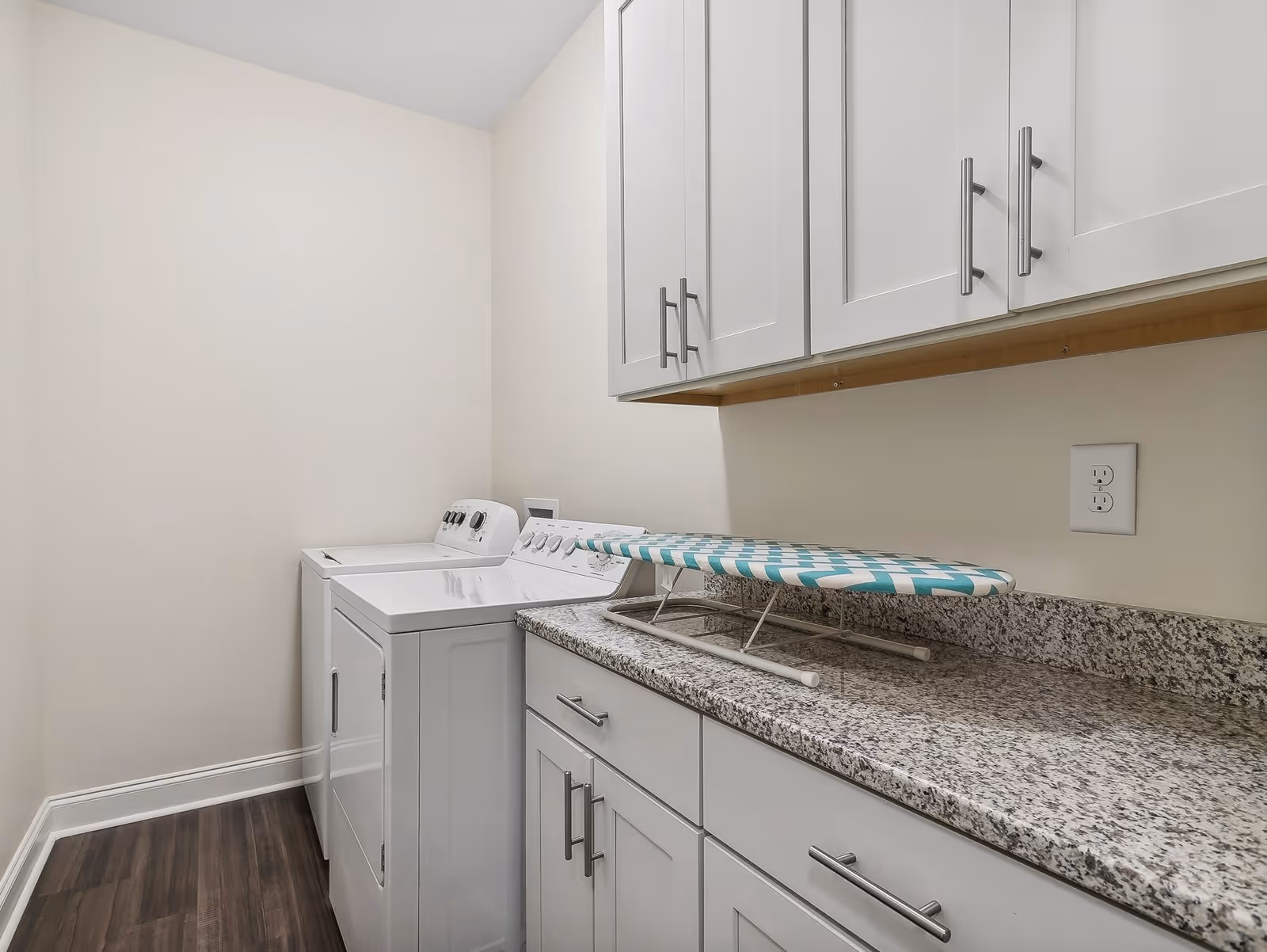 Laundry room with a white washing machine and dryer, white cabinets above and below a granite countertop, and an ironing board placed on the countertop.