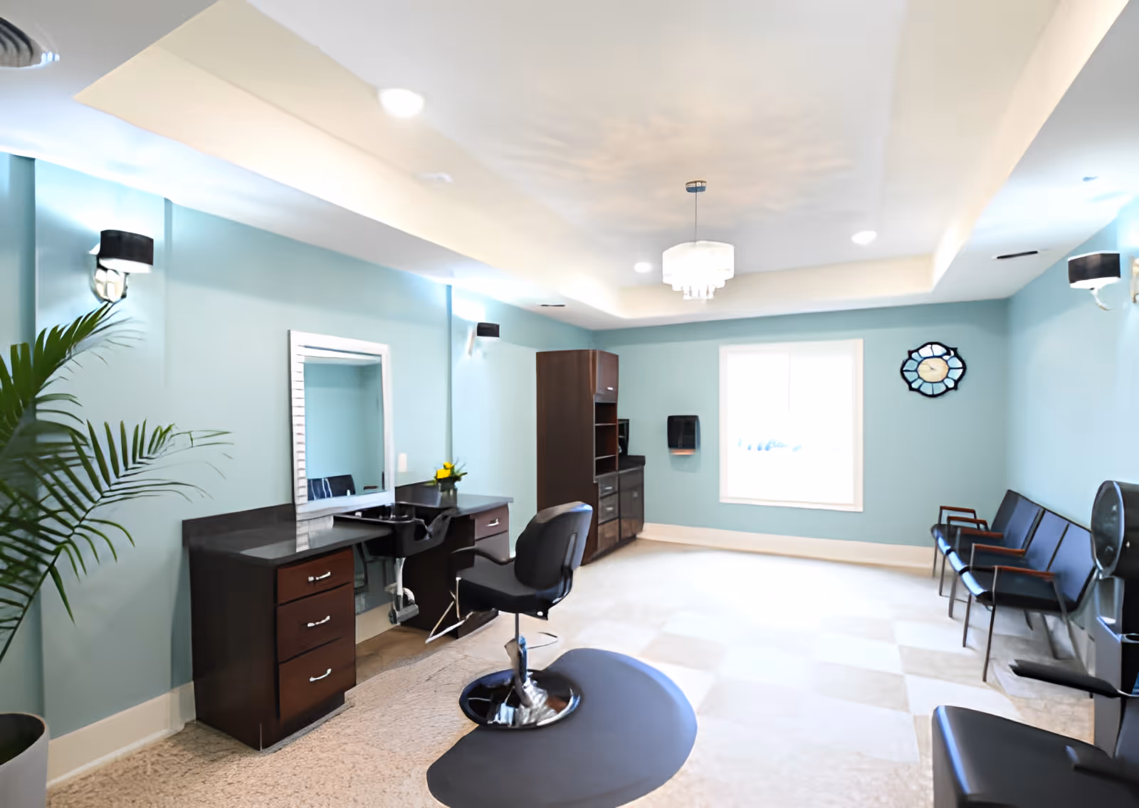A bright and clean hair salon area inside a senior living facility with light blue walls, a styling chair in front of a large mirror, dark wood cabinets, a chandelier, and seating along the opposite wall.