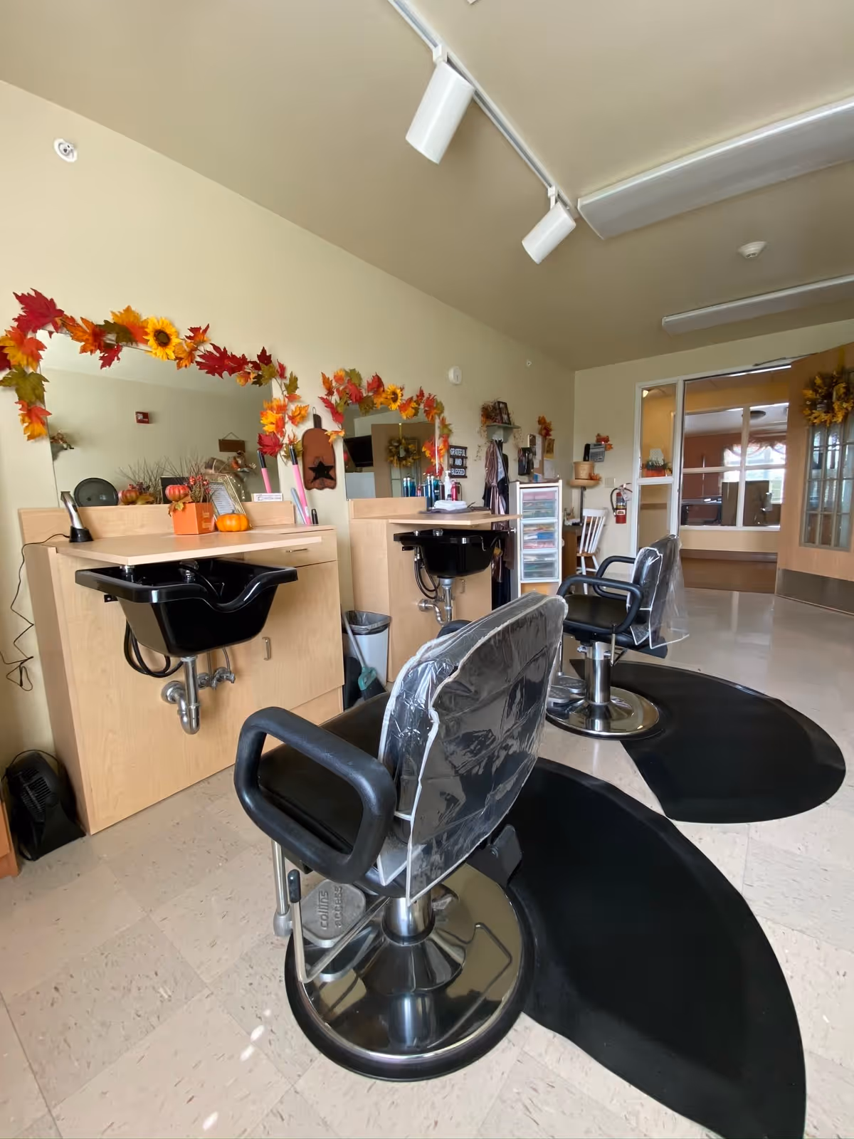 Interior view of a hair salon area in a senior living facility with two black salon chairs covered in plastic, black wash basins, large mirrors decorated with autumn leaves and sunflowers, and various hair care products on the counters. The room has beige walls and a tiled floor, with a door and windows visible in the background.