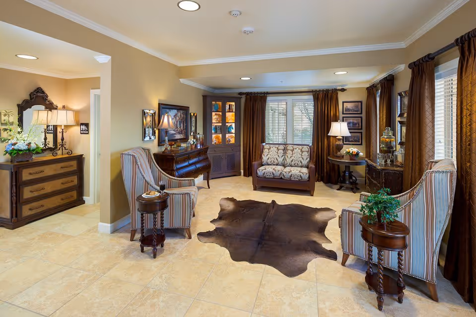 A cozy living room area in Northbrook Inn Memory Care Community featuring two striped armchairs with small round wooden side tables, a patterned loveseat, a dark brown cowhide rug on tiled floor, and warm beige walls. The room is decorated with framed artwork, a wooden dresser with a lamp and flowers, a corner cabinet with decorative items, and large windows with brown curtains allowing natural light to enter.