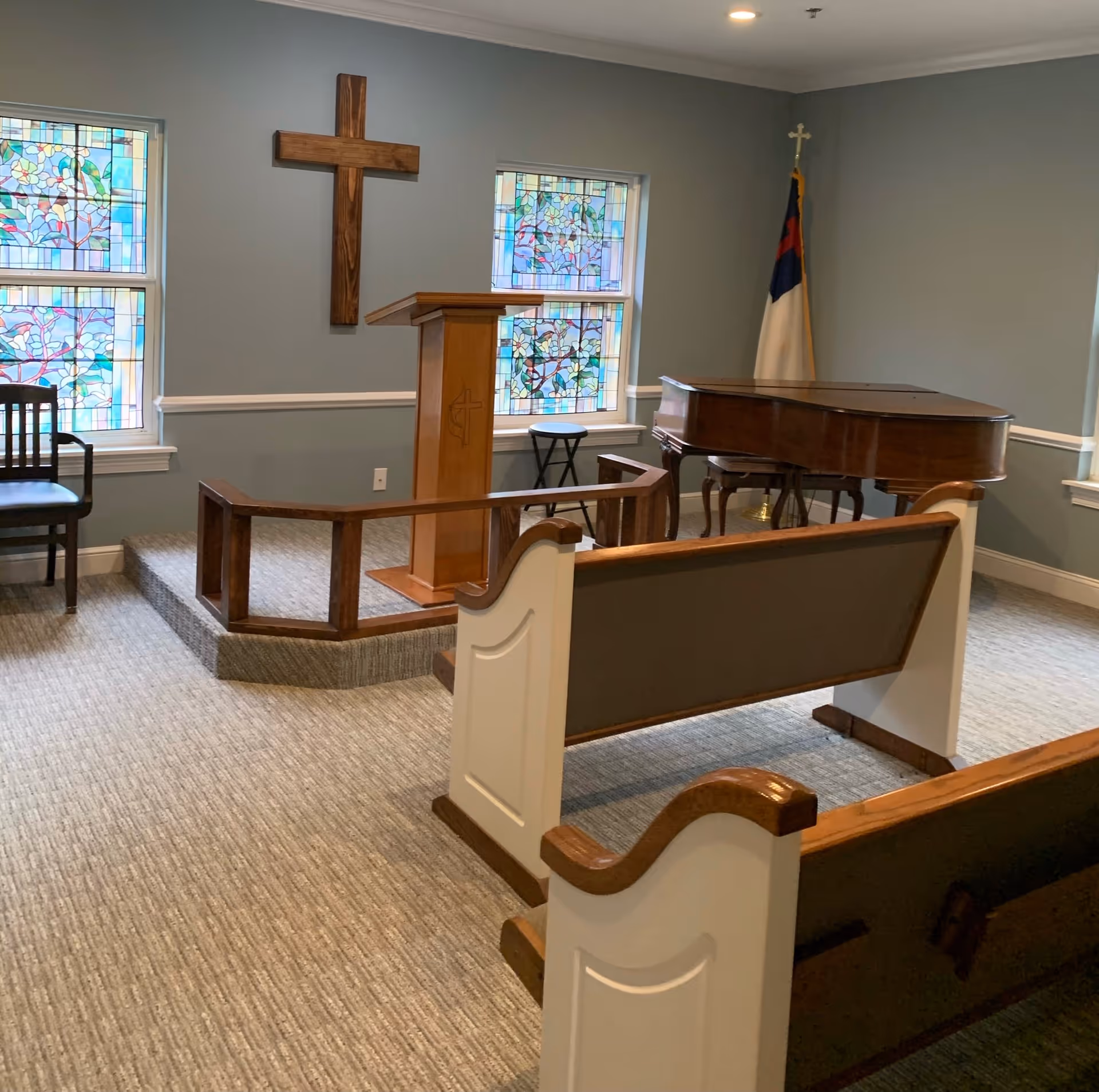 Interior of a small chapel room with wooden pews, a wooden podium with a cross symbol, a large wooden cross mounted on the wall, stained glass windows with floral designs, a grand piano, and a Christian flag in the corner.